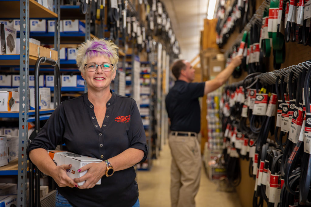 woman holding an auto part in front of shelves of auto parts with a man picking a part from the shelf in the background